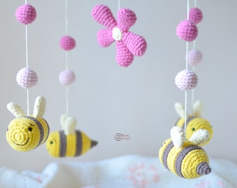 May include: A pink and yellow crocheted baby mobile with bees, flowers, and balls hanging from a white ceiling.