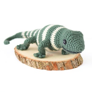 May include: A green and white striped crocheted lizard with black eyes. The lizard is sitting on a wooden slice.