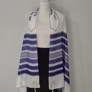 Op de afbeelding: Witte tallit met blauwe en paarse strepen, een zilveren Davidster aan elke kant en Hebreeuwse tekst bovenaan. De tallit is over een wit mannequin gehangen.