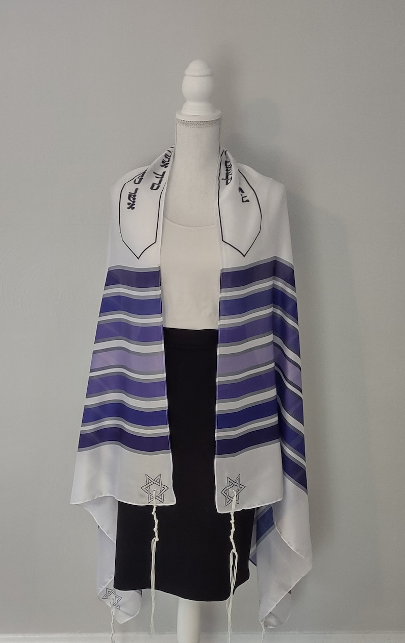 Op de afbeelding: Witte tallit met blauwe en paarse strepen, een zilveren Davidster aan elke kant en Hebreeuwse tekst bovenaan. De tallit is over een wit mannequin gehangen.