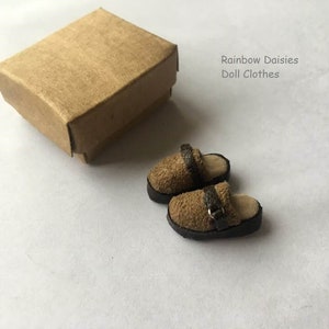 Könnte beinhalten: Ein Paar braune Wildlederschuhe für Puppen mit schwarzen Schnallen. Die Schuhe liegen auf einer weißen Oberfläche neben einem braunen Karton. Der Text "Rainbow Daisies Doll Clothes" ist in der oberen rechten Ecke des Bildes zu sehen.