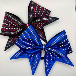Può includere: Tre fiocchi da cheerleader neri e blu. Il fiocco nero ha strass rosa e iridescenti, mentre il fiocco blu ha strass iridescenti. Entrambi i fiocchi sono impreziositi da accenti scintillanti.