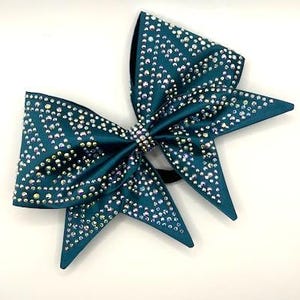 May include: A teal cheerleading bow with iridescent rhinestones. The bow is made of satin material and has a central rhinestone embellishment. The bow is designed with multiple points and is attached to a black elastic band.