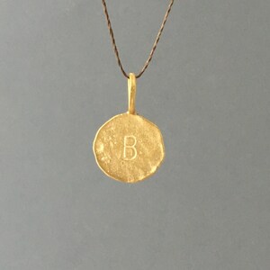 以下が含まれることがあります： 丸いチャームに「B」の文字が刻まれた、ゴールドのペンダントネックレス。チャームはテクスチャ加工が施され、ハンマーで叩かれたような仕上げになっています。
