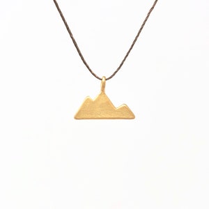 Puede incluir: Un collar con un colgante de montaña de oro en un cordón marrón.