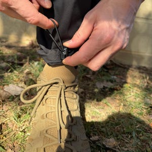 May include: Black pants with a drawstring and a black toggle closure at the bottom of the leg. The pants are being worn by a person with brown hiking boots.