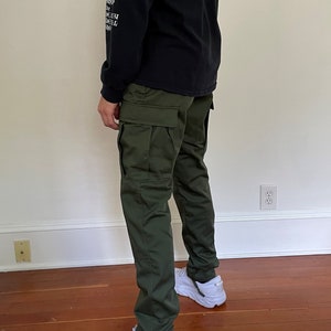 May include: A person wearing a black long-sleeved shirt with white text that says "Heavy Metal Shop" and green cargo pants. The person is wearing white sneakers with the word "Hoka" on them.
