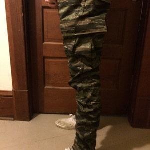May include: A pair of camouflage cargo pants with a green, brown, and black pattern. The pants are worn by a person standing in front of a wooden door.