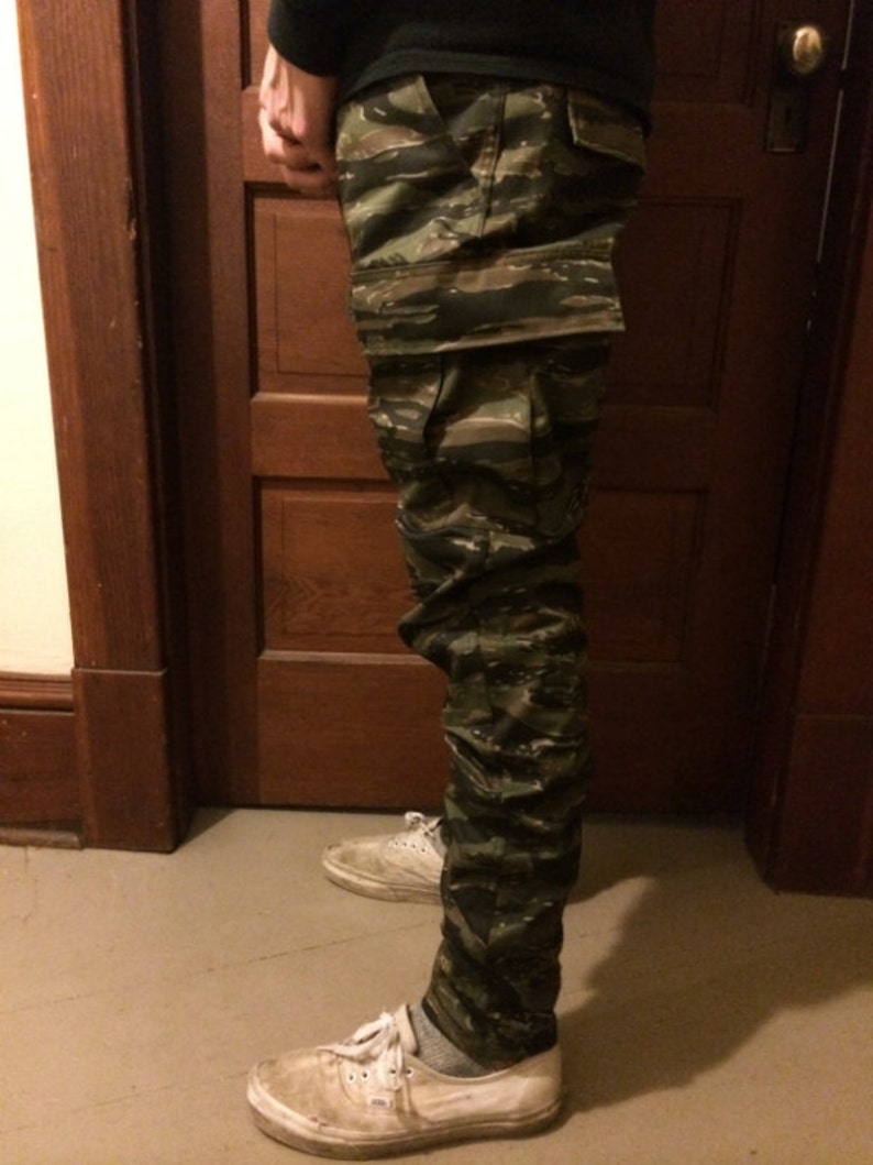May include: A pair of camouflage cargo pants with a green, brown, and black pattern. The pants are worn by a person standing in front of a wooden door.