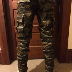 May include: A pair of camouflage cargo pants with a tiger stripe pattern. The pants have two large cargo pockets on the sides.