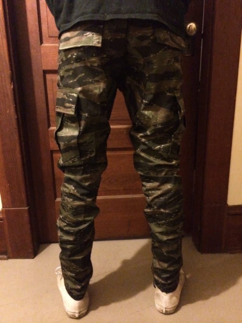 May include: A pair of camouflage cargo pants with a tiger stripe pattern. The pants have two large cargo pockets on the sides.