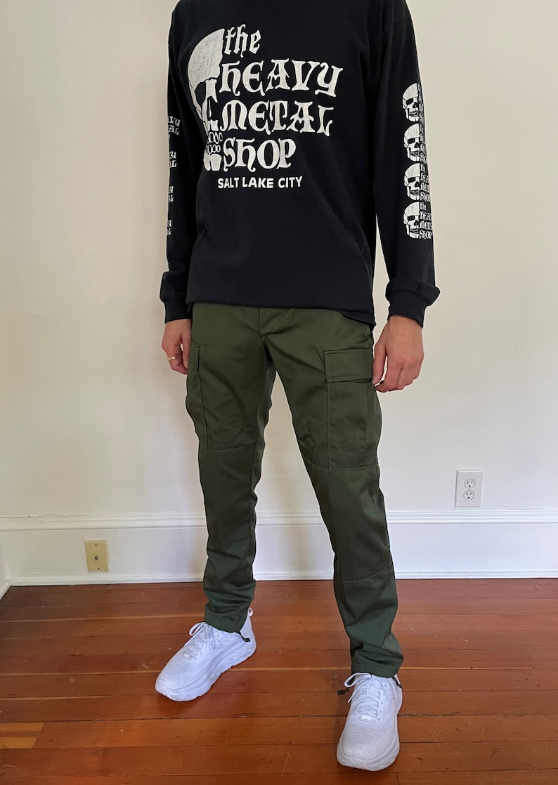 May include: A person wearing a black long-sleeved shirt with a white skull graphic and the text "The Heavy Metal Shop Salt Lake City". They are also wearing green cargo pants and white sneakers.