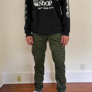 May include: A person wearing a black long-sleeve t-shirt with a white skull graphic and the text "The Heavy Metal Shop Salt Lake City". They are also wearing green cargo pants and white sneakers.