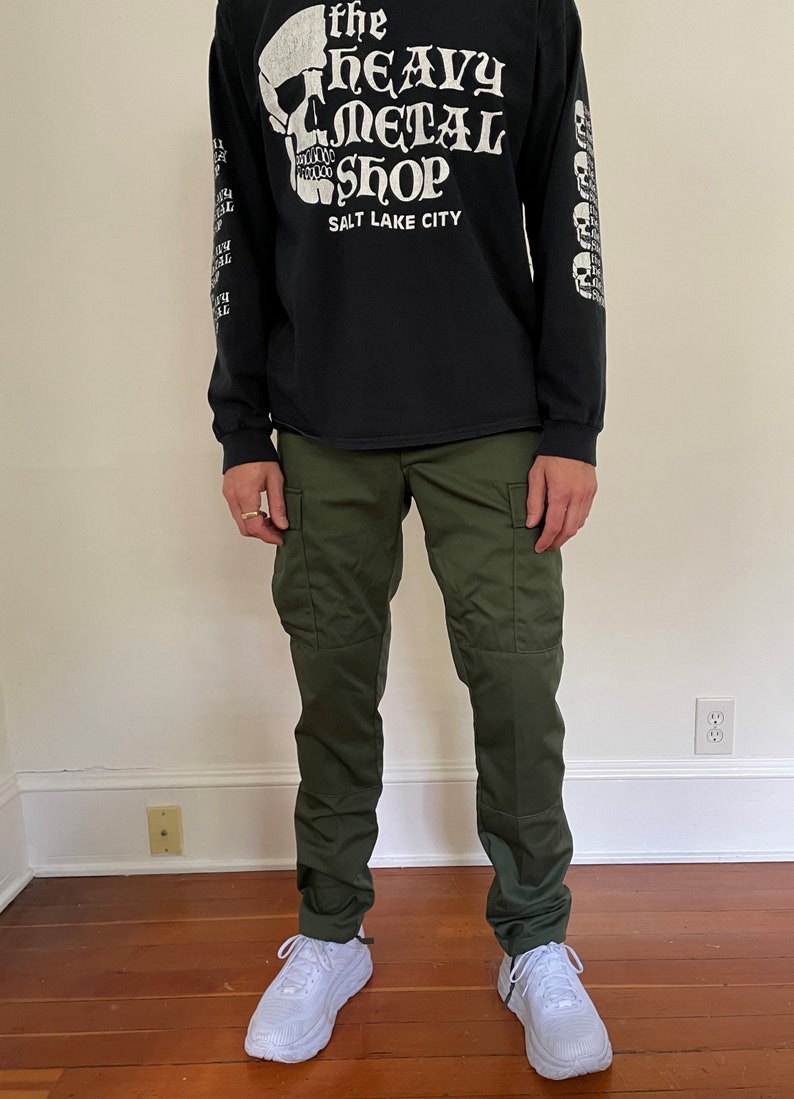 May include: A person wearing a black long-sleeve t-shirt with a white skull graphic and the text "The Heavy Metal Shop Salt Lake City". They are also wearing green cargo pants and white sneakers.