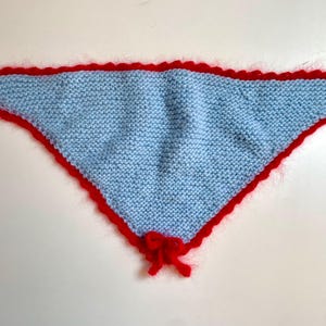 May include: A light blue knitted triangle scarf with a red scalloped edge and a red bow at the bottom.