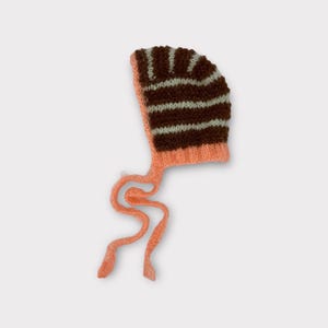 May include: A knitted baby bonnet in shades of brown, peach, and white. The bonnet features horizontal stripes on the crown and long, peach-colored ties. The soft, textured knit suggests a handmade item, perfect for a newborn.