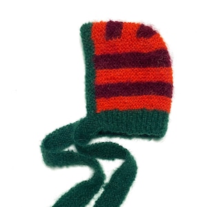 May include: A green knitted baby bonnet with a striped pattern of orange and burgundy. The bonnet has a long tie that can be tied under the chin.