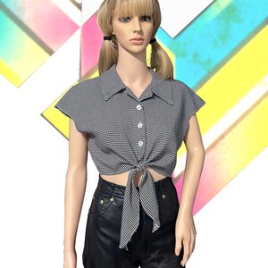 May include: A black and white gingham print short-sleeve button-down shirt with a tie-front hem, paired with black leather shorts. The shirt features a classic collar and three visible white buttons, while the shorts have gold-tone hardware.