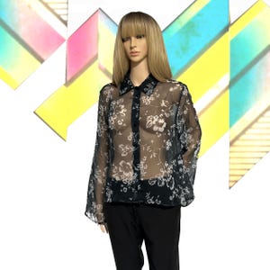 Puede incluir: Una blusa negra transparente con un estampado floral blanco y mangas largas. La blusa tiene cuello camisero y se combina con pantalones negros. El fondo presenta formas geométricas coloridas.