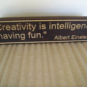 May include: A rectangular wooden sign with a dark brown finish and a lighter brown border. The sign features the quote "Creativity is intelligence having fun." attributed to Albert Einstein.
