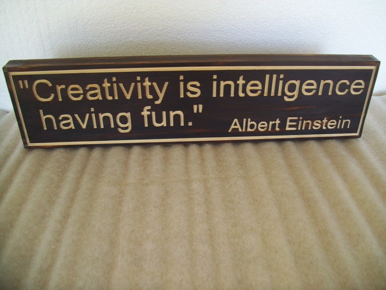 May include: A rectangular wooden sign with a dark brown finish and a lighter brown border. The sign features the quote "Creativity is intelligence having fun." attributed to Albert Einstein.