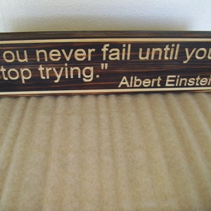 May include: A rectangular wooden sign with the quote "You never fail until you stop trying." attributed to Albert Einstein. The sign has a dark brown center with lighter wood trim and lettering.
