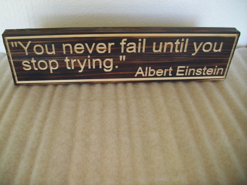 May include: A rectangular wooden sign with the quote "You never fail until you stop trying." attributed to Albert Einstein. The sign has a dark brown center with lighter wood trim and lettering.