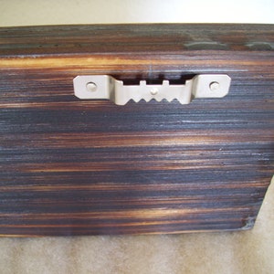 May include: Close-up of a wooden plank with a dark, burnt finish and visible wood grain. A metal sawtooth hanger is attached to the back, ready for wall mounting. The wood has a rustic, textured appearance.