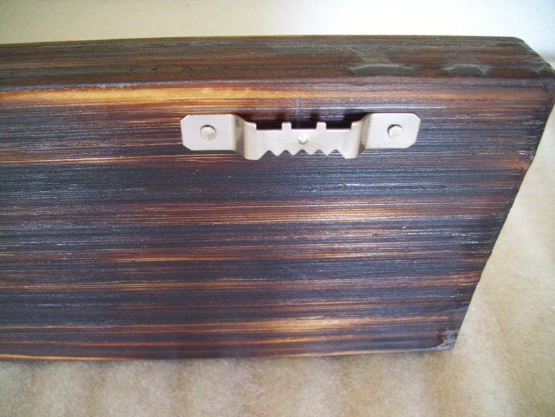 May include: Close-up of a wooden plank with a dark, burnt finish and visible wood grain. A metal sawtooth hanger is attached to the back, ready for wall mounting. The wood has a rustic, textured appearance.