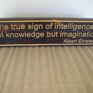 May include: A rectangular wooden sign with the quote "The true sign of intelligence is not knowledge but imagination. Albert Einstein" engraved in a light color. The wood has a dark, burnt appearance.