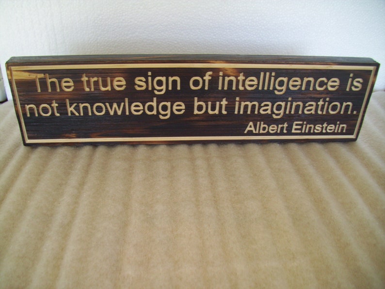 May include: A rectangular wooden sign with the quote "The true sign of intelligence is not knowledge but imagination. Albert Einstein" engraved in a light color. The wood has a dark, burnt appearance.