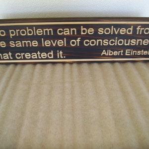 May include: A rectangular wooden sign with the quote "No problem can be solved from the same level of consciousness that created it." by Albert Einstein. The text is in a light color against a dark wood background.