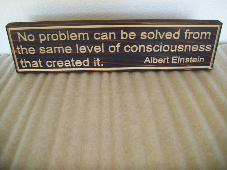 May include: A rectangular wooden sign with the quote "No problem can be solved from the same level of consciousness that created it." by Albert Einstein. The text is in a light color against a dark wood background.
