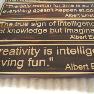 May include: Wooden plaques with quotes by Albert Einstein. The plaques have a dark brown background with gold-colored lettering and a gold border. The quotes include: "The only reason for time is so that everything doesn't happen at once."