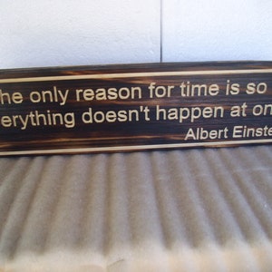 May include: A rectangular wooden sign with a dark brown finish and a lighter border. The sign features the quote "The only reason for time is so that everything doesn't happen at once." by Albert Einstein, engraved in a light color.
