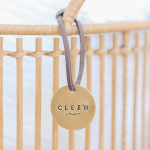 Pode incluir: Uma etiqueta redonda dourada com a palavra "CLEAN" estampada em preto, pendurada em uma cesta de vime. A etiqueta é suspensa por uma fina tira de couro cinza. A cesta é feita de material tecido marrom claro.