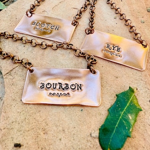 May include: Three copper-colored metal liquor tags with chain attachments. The tags are rectangular and stamped with the words "SCOTCH", "RYE", and "BOURBON" in a decorative font. A green leaf rests nearby.