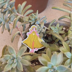May include: A sticker of a cartoon character with pink hair, a yellow cloak, and a star. The sticker is placed among green succulent plants in a grey pot, with a brown pot in the background.