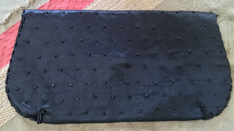 Puede incluir: Un bolso de noche negro con cuentas, con una textura lisa y satinada. El bolso est&aacute; adornado con peque&ntilde;as cuentas negras que est&aacute;n espaciadas uniformemente por toda la superficie.
