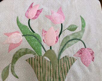 May include: A quilted wall hanging featuring a bouquet of pink tulips with green leaves and stems. The tulips are arranged in a green and white striped pot.