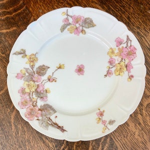 May include: A white porcelain plate with a scalloped edge features a delicate floral design. The design includes pink and yellow flowers with green leaves, arranged in a symmetrical pattern around the plate.