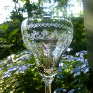 May include: A clear glass wine glass with a decorative etched design around the rim. The glass has a fluted design and a long stem.