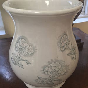 May include: A white ceramic vase with a wide mouth and a bulbous body. The vase is decorated with a green floral pattern.