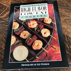 May include: A black cookbook with the title "HIGH-FLAVOR LOW-FAT COOKING" by Steven Raichlen. The cover features a photo of shrimp and meat skewers on a black plate. The book has won a James Beard Award.