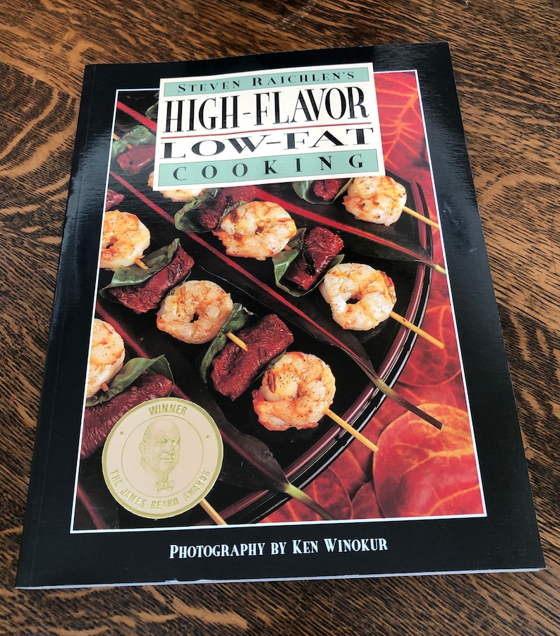 May include: A black cookbook with the title "HIGH-FLAVOR LOW-FAT COOKING" by Steven Raichlen. The cover features a photo of shrimp and meat skewers on a black plate. The book has won a James Beard Award.
