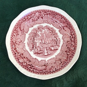 May include: A vintage pink and white ceramic saucer with a detailed floral design featuring a central image of a building and figures in a garden setting.