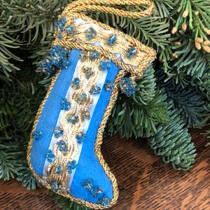 May include: A blue felt stocking ornament with gold trim and blue beads. The stocking has a white ribbon with gold trim.