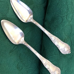 May include: Two silver spoons with ornate handles featuring a floral design. The spoons have a pointed, rounded shape.