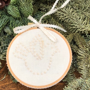 May include: A white fabric ornament with a simple design of white beads stitched in the shape of a flower. The ornament is in a wooden embroidery hoop and has a white satin ribbon attached.