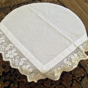 May include: White round tablecloth with a delicate white crocheted lace border. The tablecloth is made of a smooth, linen-like fabric.